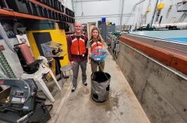 两名主要研究学生研究人员将他们赢得的海洋可再生能源设备放在波浪箱前. 他们穿着红色的救生衣.